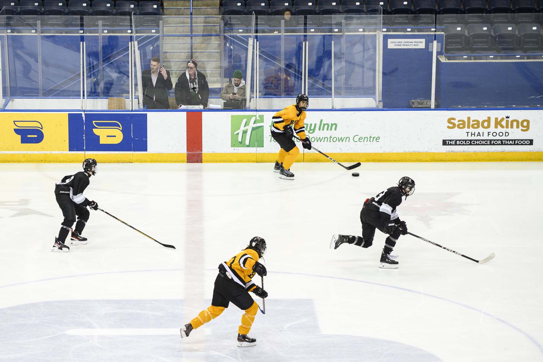 Players from opposing teams move the puck forward across the centre line while bystanders look on from the stands.